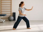 Persona in piedi in una sala per attività motorie che esegue un movimento lento e controllato con le braccia, ispirato alle pratiche di Tai Chi e Qi Gong. Sullo sfondo sono visibili una spalliera in legno, tappetini e attrezzature per l'esercizio fisico.